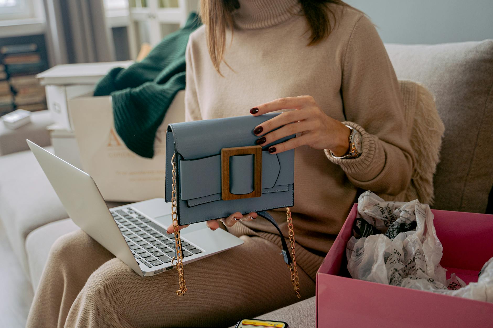 Kobieta na sofie rozpakowuje torebkę kupioną online przy użyciu laptopa