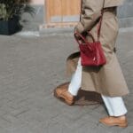 Kobieta w beżowym trenczu idzie chodnikiem z czerwoną torebką