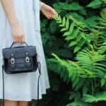 Kobieta w pasiistej sukience trzyma czarną torebkę przy zielonych roślinach